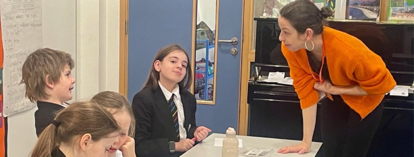 Poet Kate Wakeling running a poetry workshop in a classroom. She's wearing an orange jumper and talking to three children, in school uniform who are sat at a desk.
