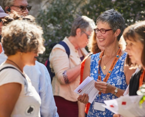 A group of people stood together with leaflets chatting in the garden of Southwell Minster. It's a sunny day, they are smiling, wearing lanyards and holding leaflets.
