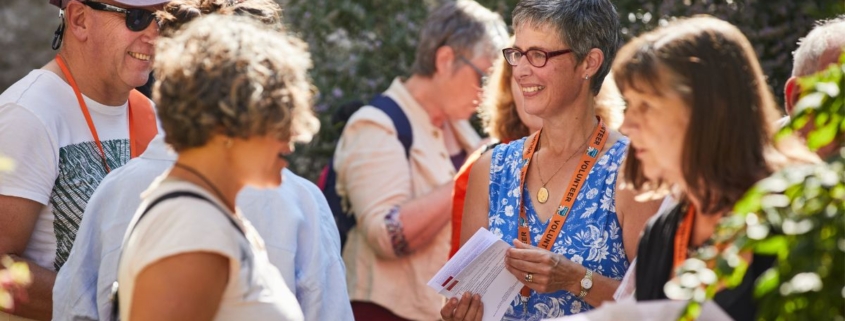 A group of people stood together with leaflets chatting in the garden of Southwell Minster. It's a sunny day, they are smiling, wearing lanyards and holding leaflets.