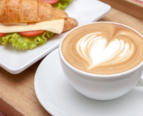 A coffee in a white cup and saucer with a heart in milk forth. It is placed next to a filled croissant with cheese and salad.