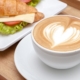 A coffee in a white cup and saucer with a heart in milk forth. It is placed next to a filled croissant with cheese and salad.