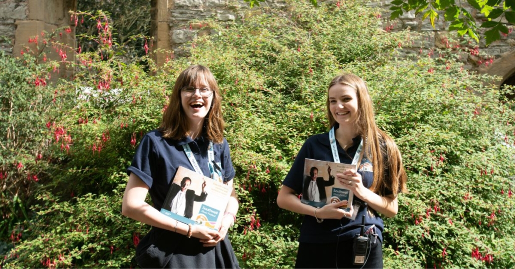 Two festival front of house volunteers stood outside, smiling together. They wear black, with lanyards around their necks and are holding a festival programme.