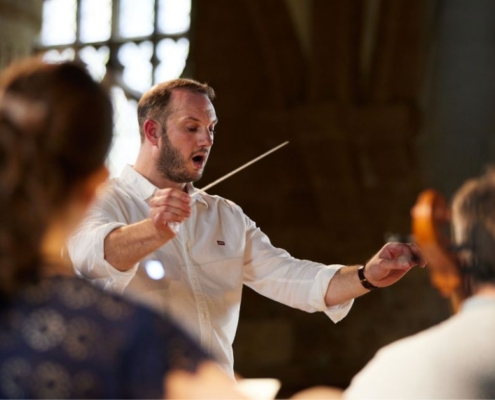 Image focuses on Marcus Farnsworth. He is wearing a casual white shirt and holds a conductor's baton in the air.