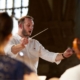 Image focuses on Marcus Farnsworth. He is wearing a casual white shirt and holds a conductor's baton in the air.