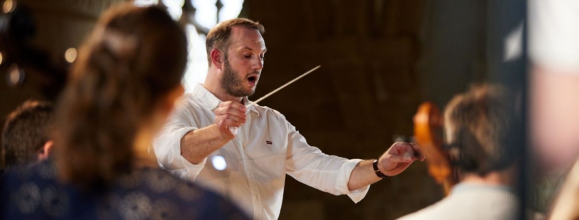 Image focuses on Marcus Farnsworth. He is wearing a casual white shirt and holds a conductor's baton in the air.