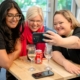 Three people gathered around a table in the Festival Marquee. There are drinks on the table and they are smiling as one of them takes a selfie on a smartphone.