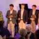 Five young performers stood together in front of an audience with a piano behind them.