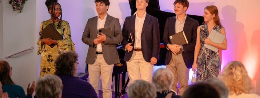Five young performers stood together in front of an audience with a piano behind them.